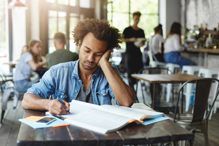 sad unhappy dark skinned bearded student feeling frustrated while preparing lessons college writing down his copybook with pen leaning elbow looking notes with upset expression